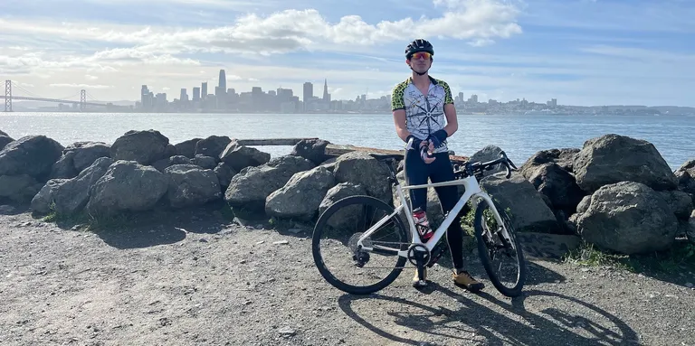 A person standing with a bike in front of the San Francisco skyline, wearing spandex and looking a little too serious and important